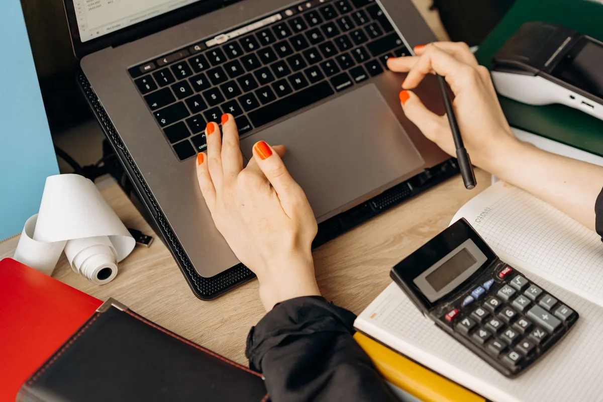 Hands on a laptop trackpad beside a calculator, an open notebook, and a receipt paper roll on an online seller's desk